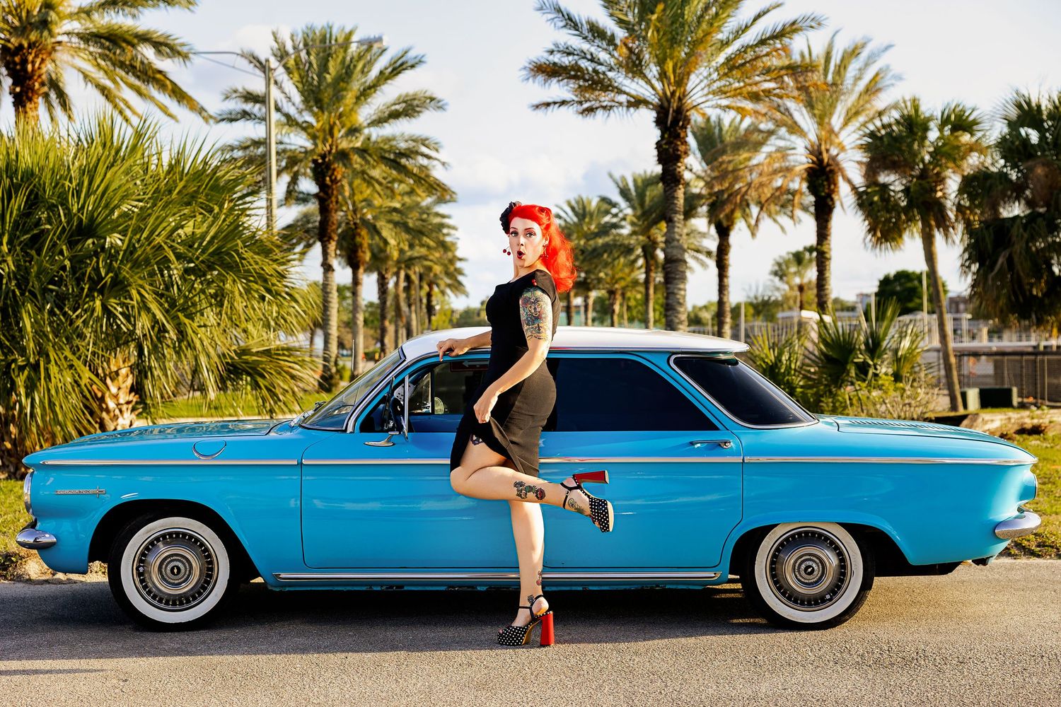 Tattooed redhead pinup woman posing beside blue classic car with palm trees background.
