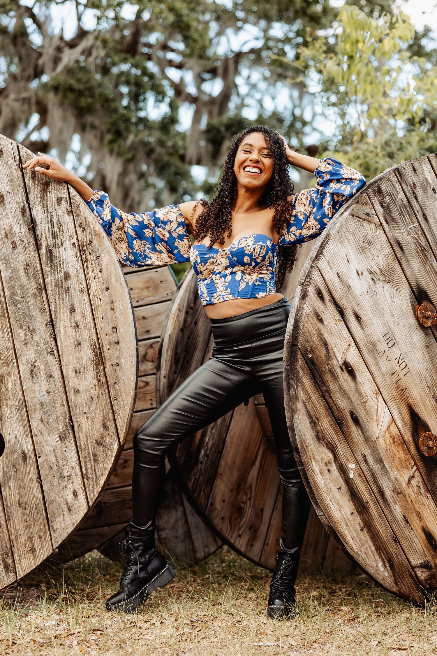 Smiling woman in blue floral top and leather pants poses between large wooden cable spools outdoors.