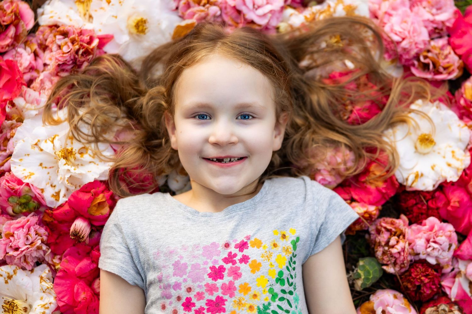 Smiling young girl with curly red hair lying among colorful pink and white roses.