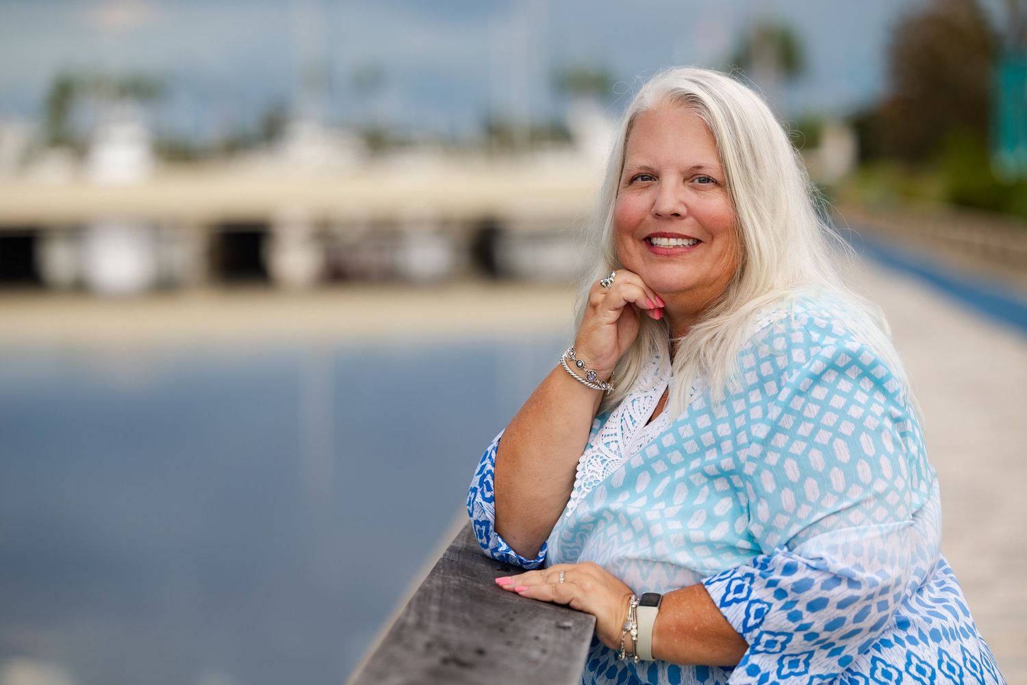 Smiling woman with long white hair leans on railing by marina waterfront wearing blue patterned top.
