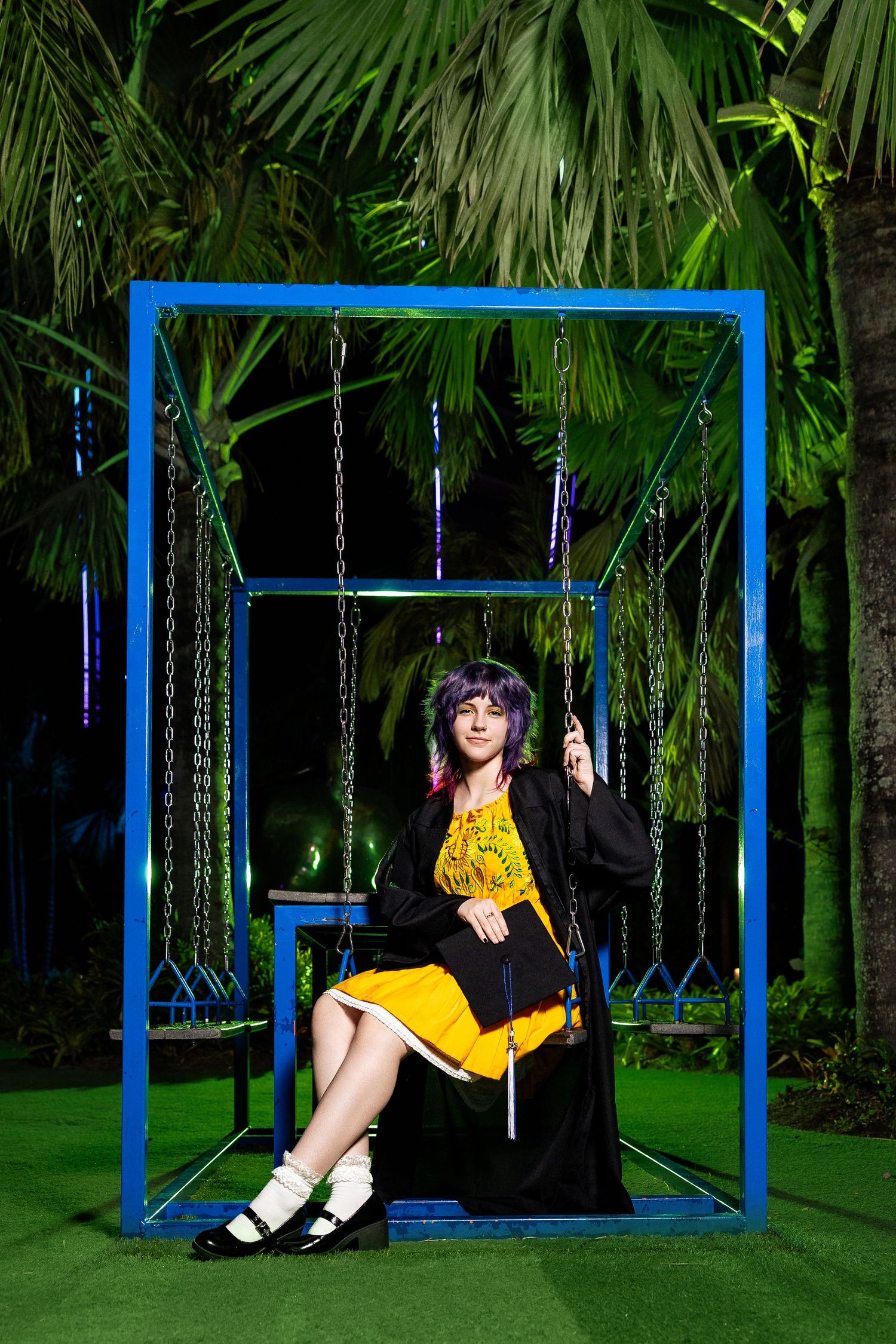 Woman with purple hair in yellow dress sits on a swing inside a blue frame structure at night.