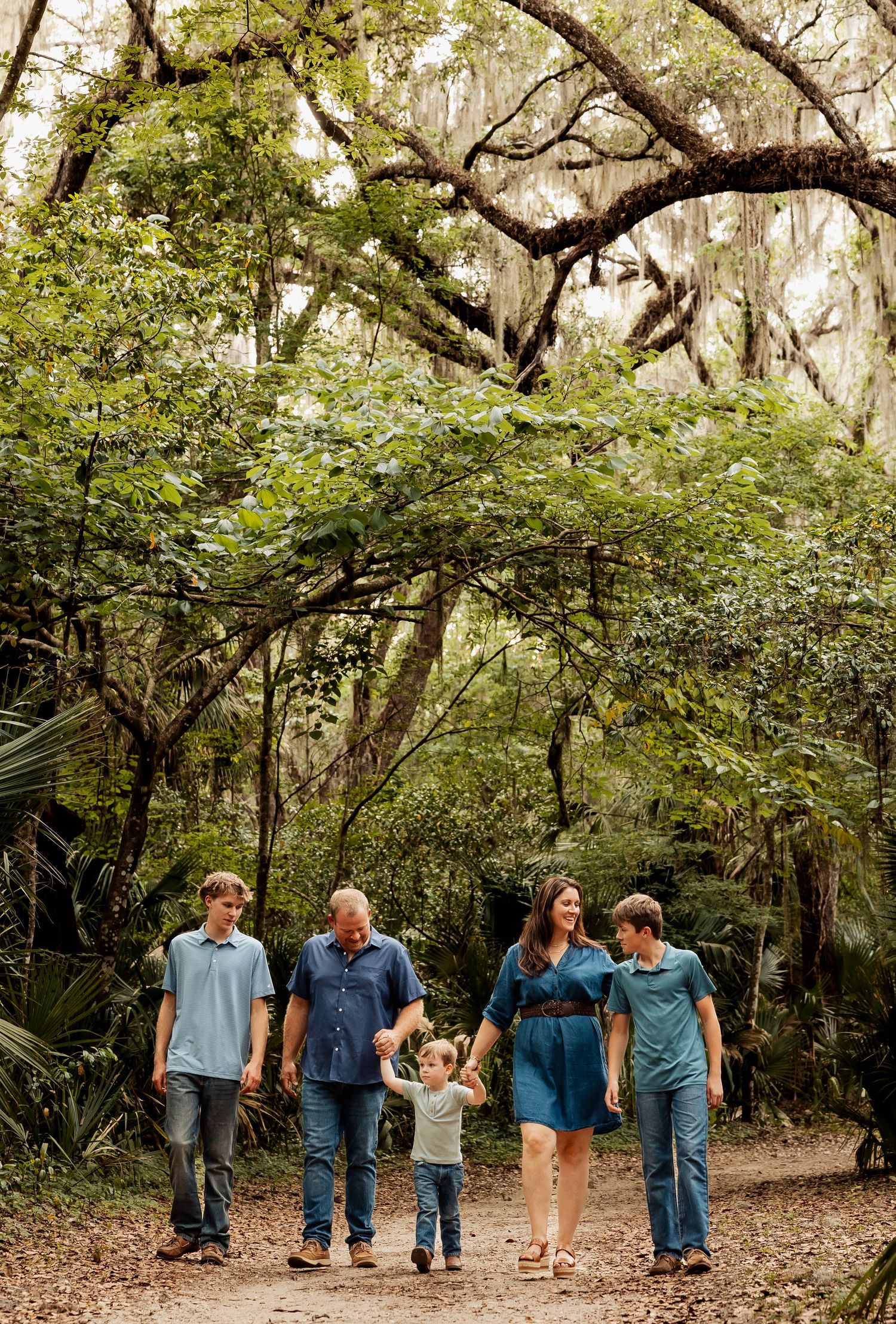 Family of five walking hand-in-hand down a shaded forest path beneath sprawling moss-covered oak trees.