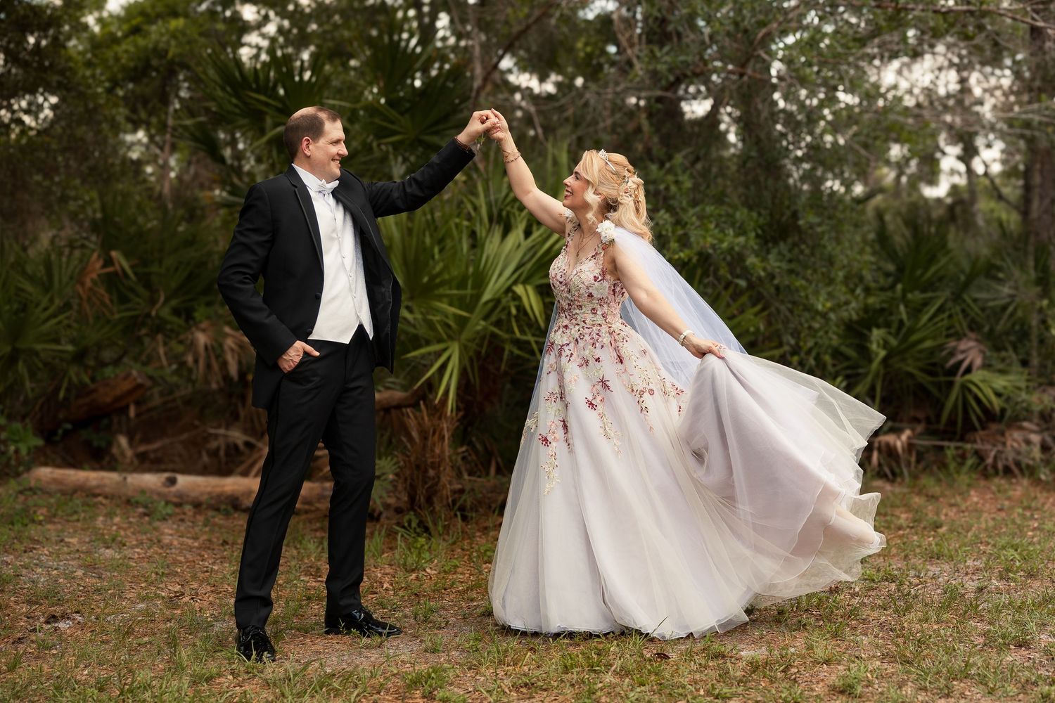 Newlyweds dancing joyfully outdoors, bride in floral tulle gown with flowing veil among tropical greenery.