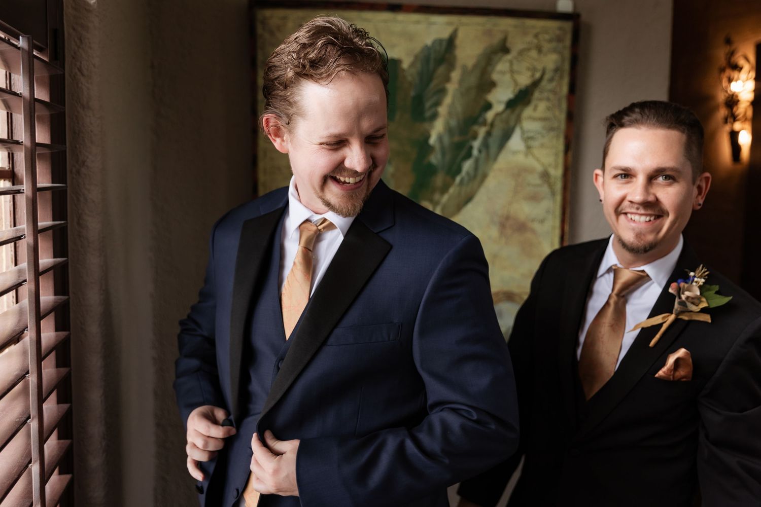 Two groomsmen in navy suits with gold ties laughing together during wedding day preparations.