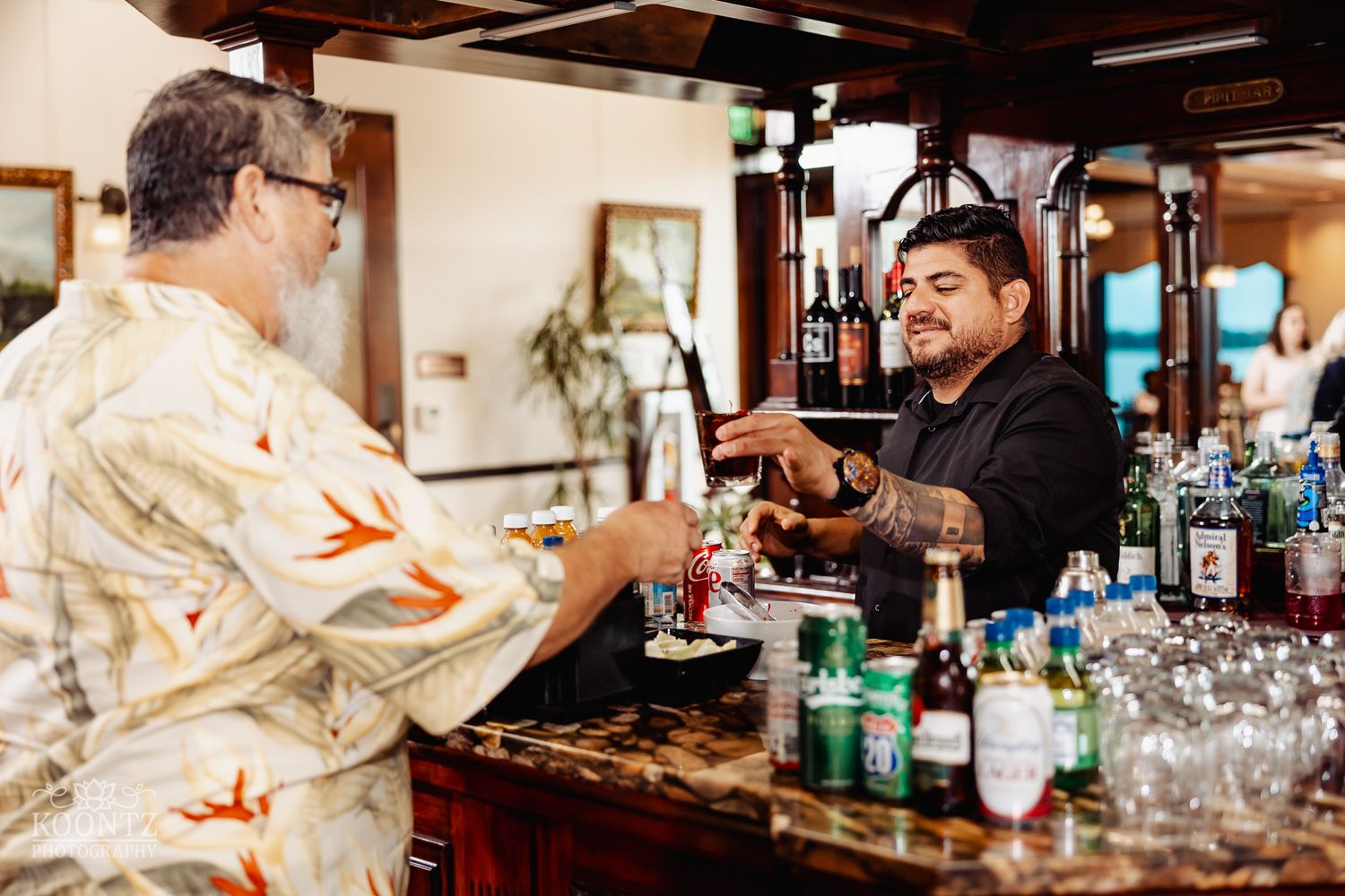 Bartender in black serving drinks to guest in Hawaiian shirt at wooden bar counter.