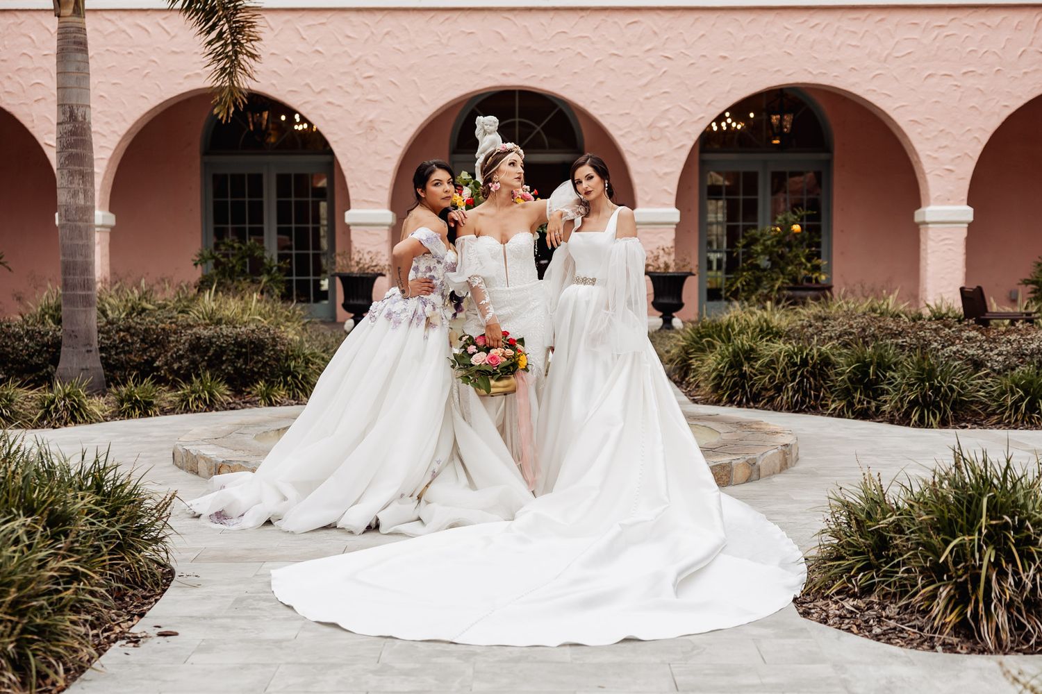 Three people in elegant white wedding gowns pose together on a pink-arched walkway with tropical landscaping.