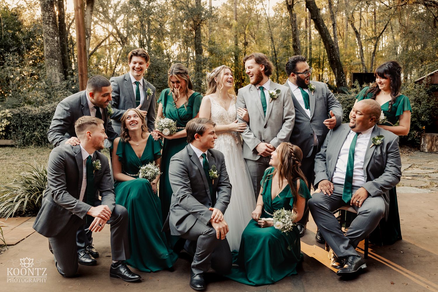 Wedding party laughs together outdoors in emerald green and gray attire at a rustic woodland wedding venue.