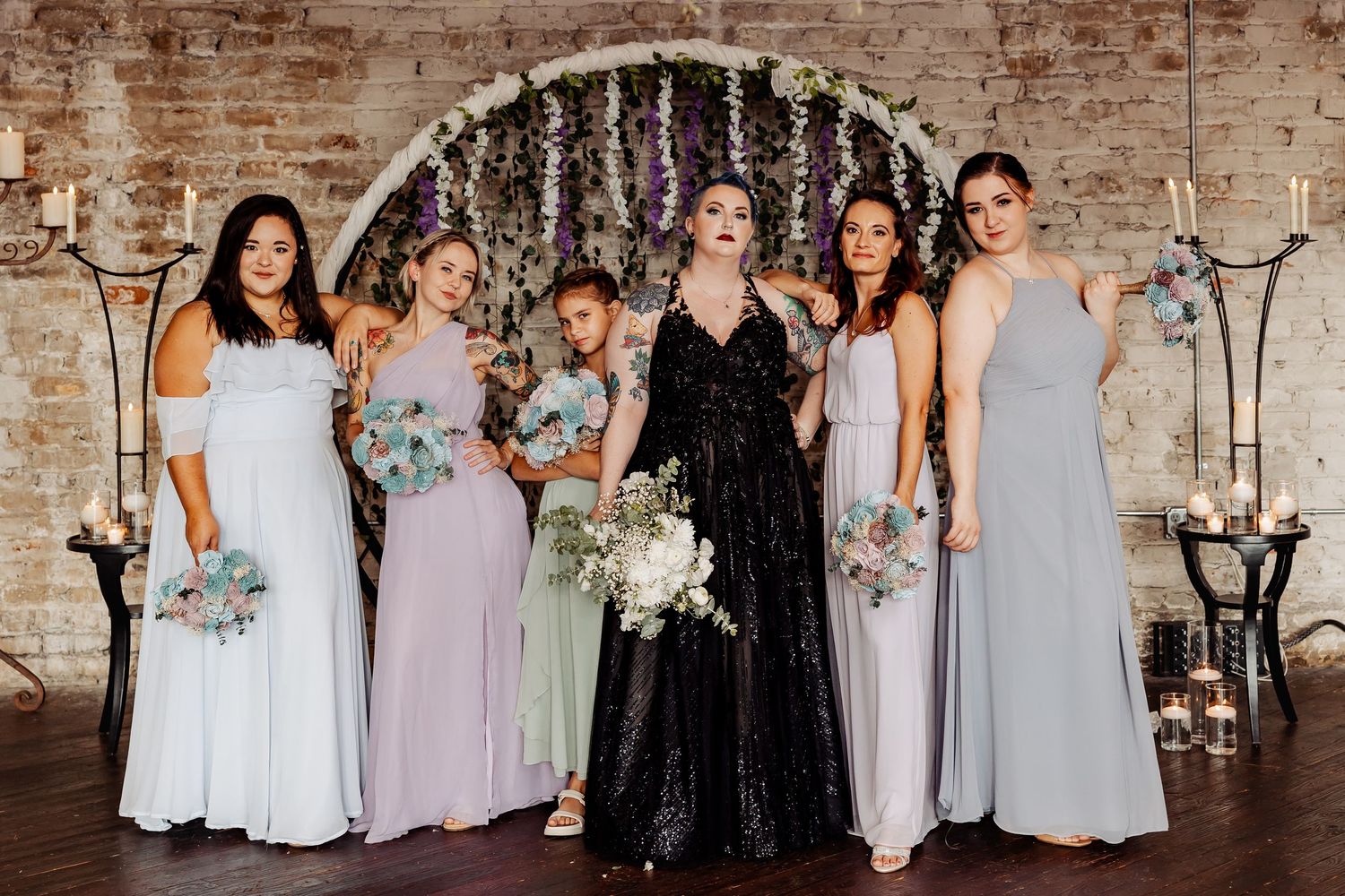 Bride in black sequin gown with bridesmaids in lavender dresses before floral circular arch.