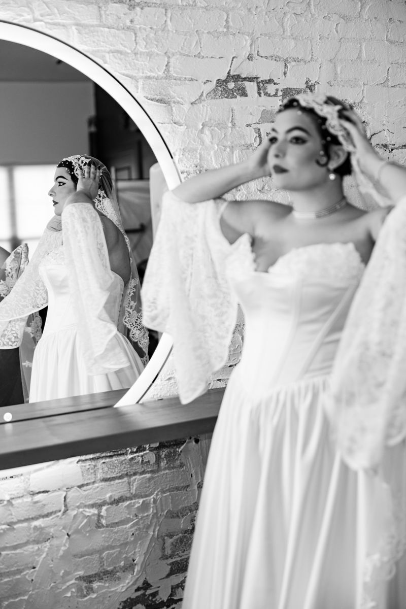 Black and white photo of bride in strapless gown adjusting lace veil in round mirror.