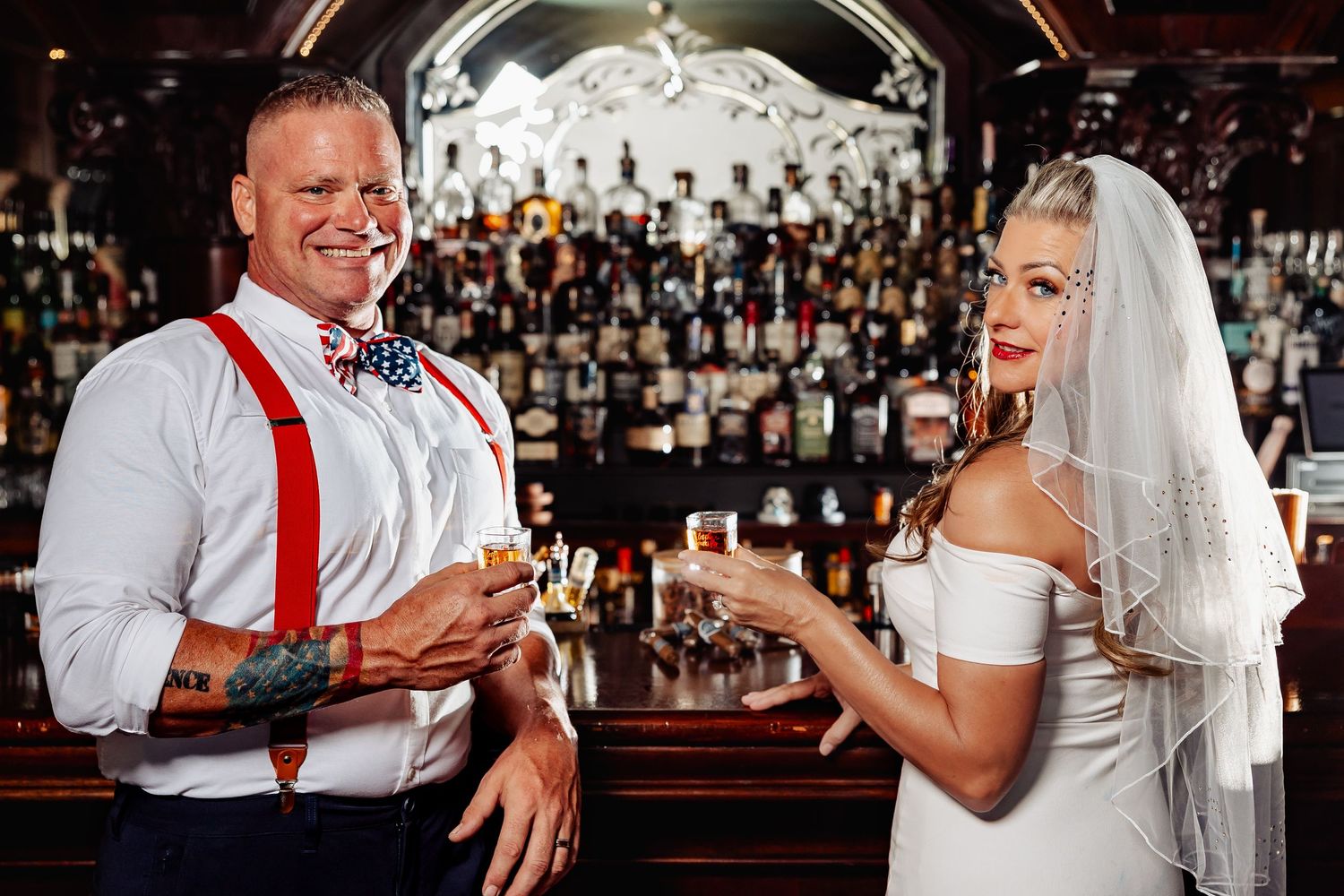Bride and groom sharing a toast at elegant bar with extensive bottle collection in background.
