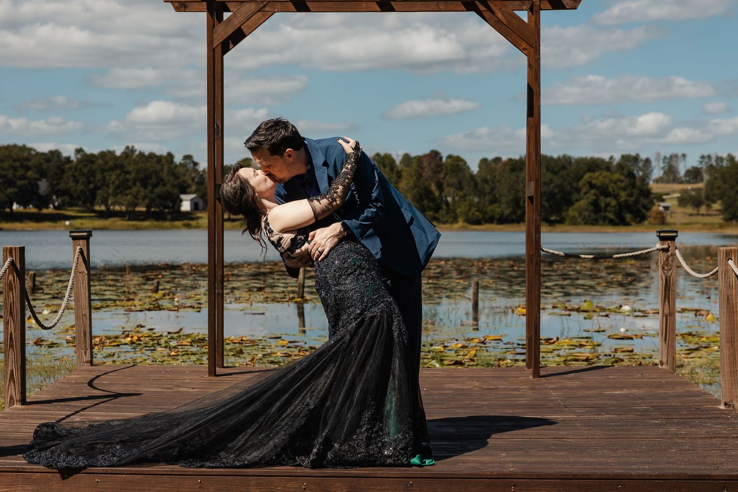 Bride in black mermaid gown and groom in blue suit sharing a dip kiss on a wooden dock by a lake.