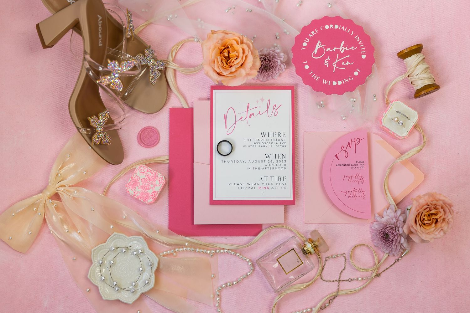 Wedding invitation flatlay with pink decor elements and florals on rosy background.