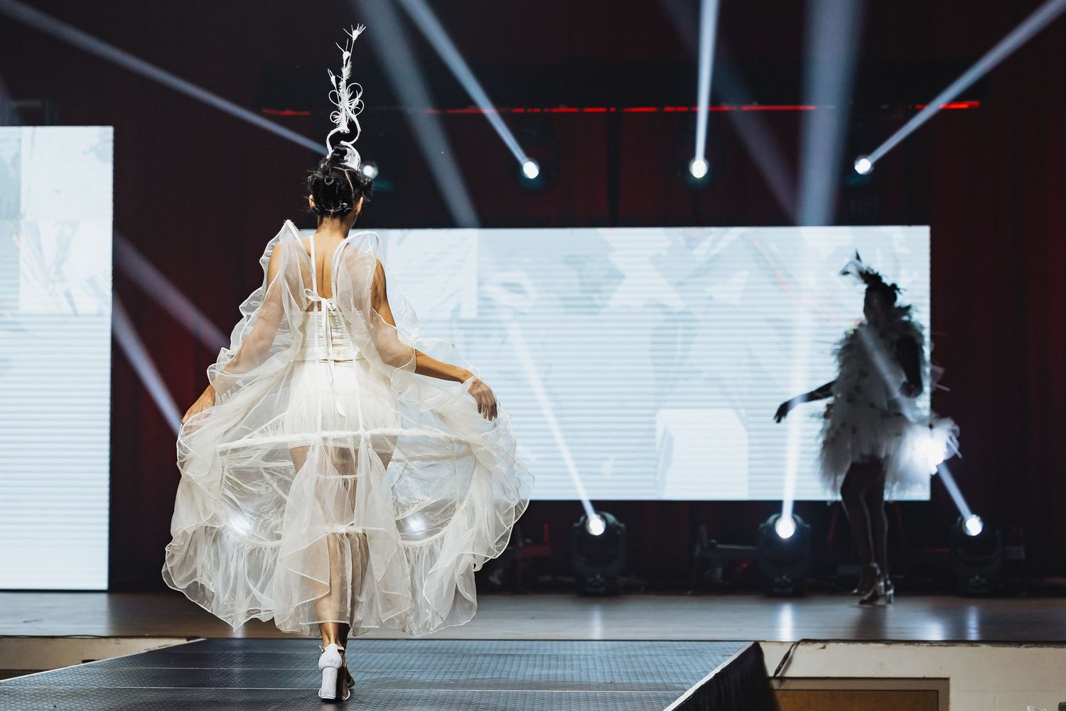 Model walks runway in flowing white tulle gown with feathered headpiece under dramatic stage lighting.