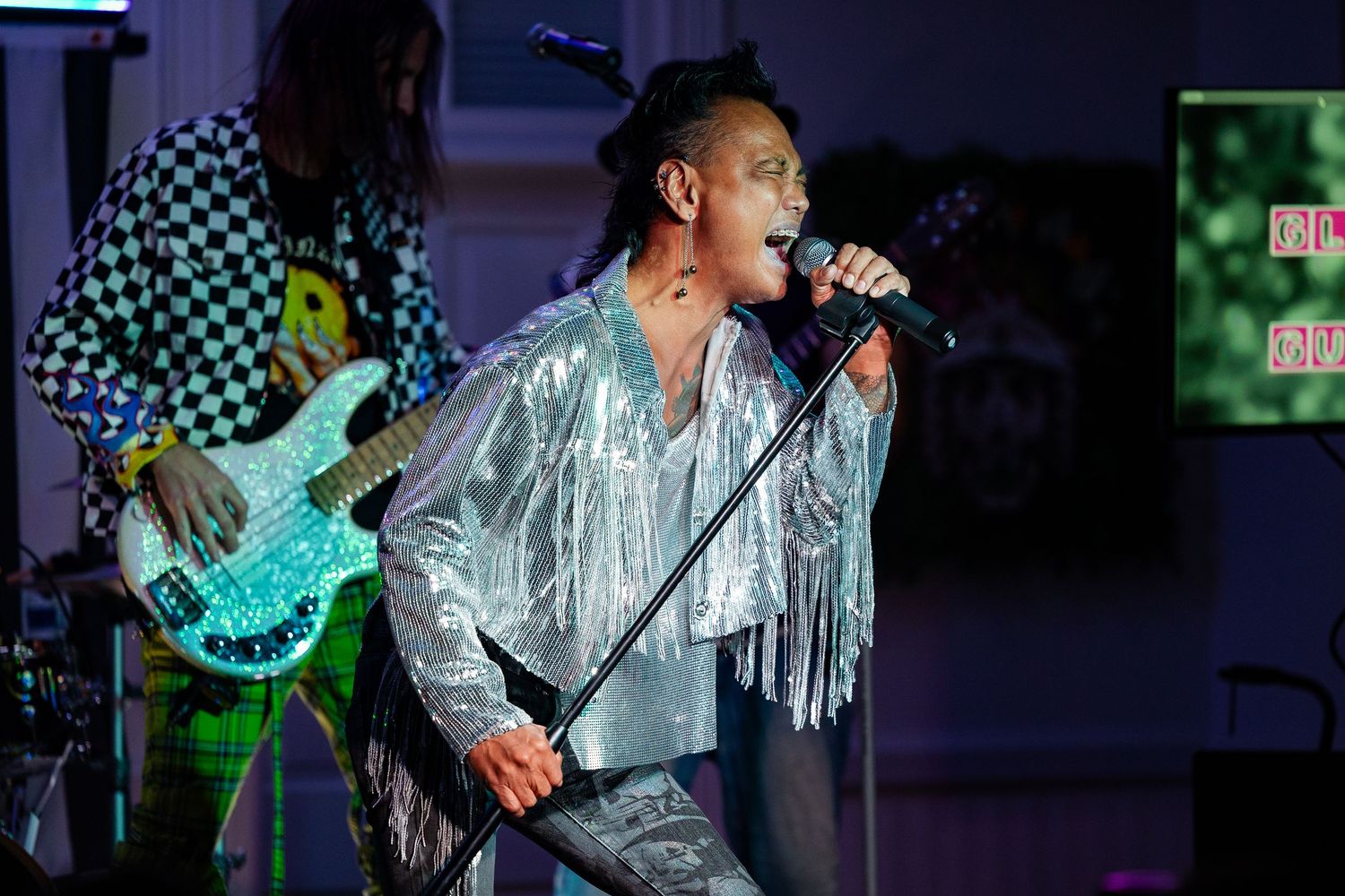 Singer in silver fringe jacket performs on stage with bassist in checkered outfit behind.