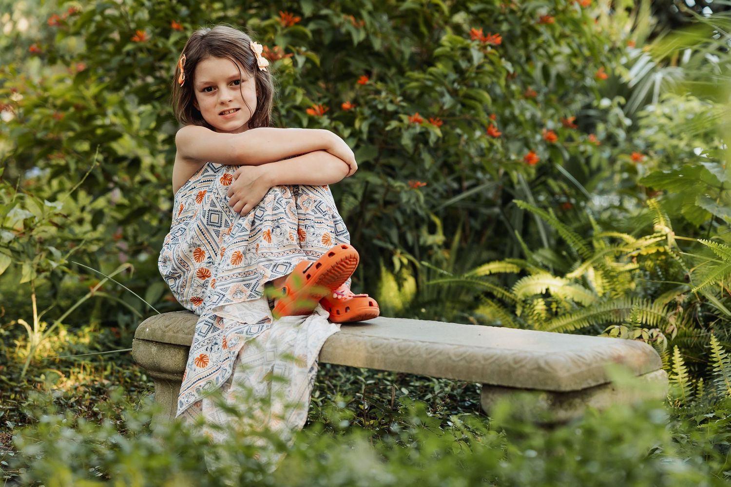A young girl in a floral outfit sits on a stone bench surrounded by lush green garden foliage.