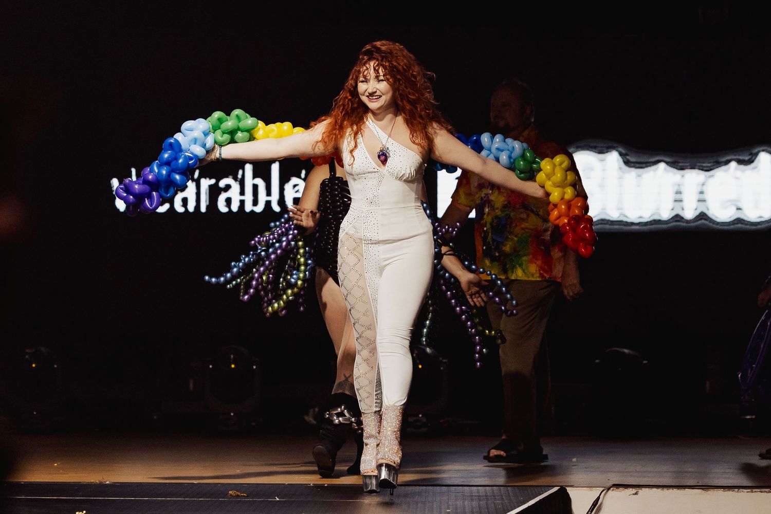 Performer in white outfit walks runway at wearable art fashion show with rainbow balloon arch backdrop.