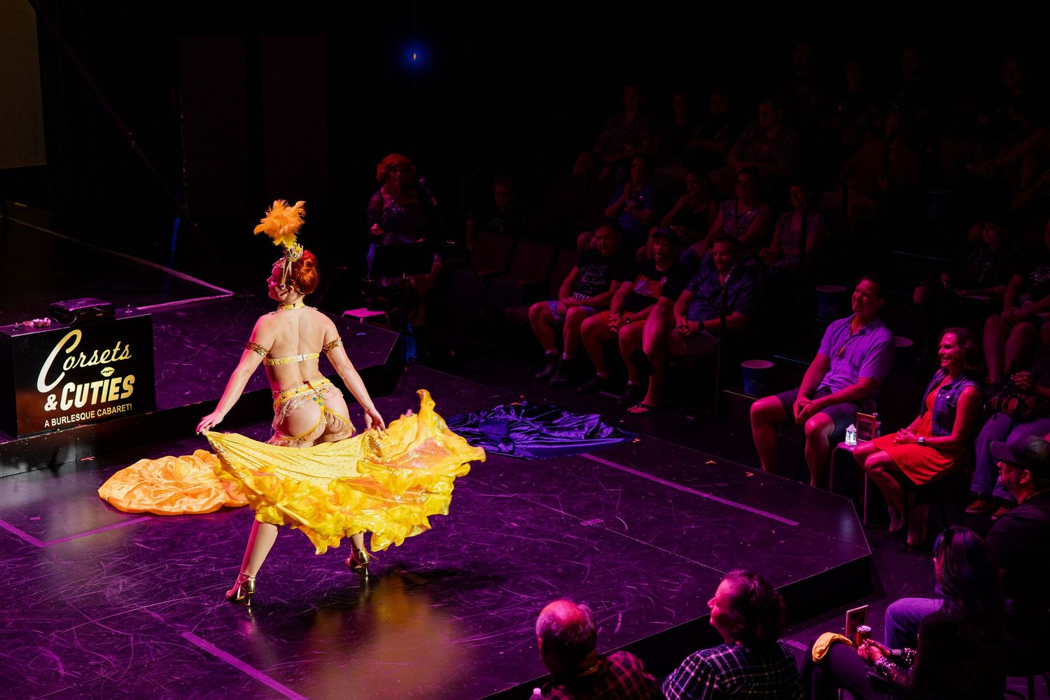 Burlesque performer in yellow ruffled costume dancing on runway stage before seated audience.