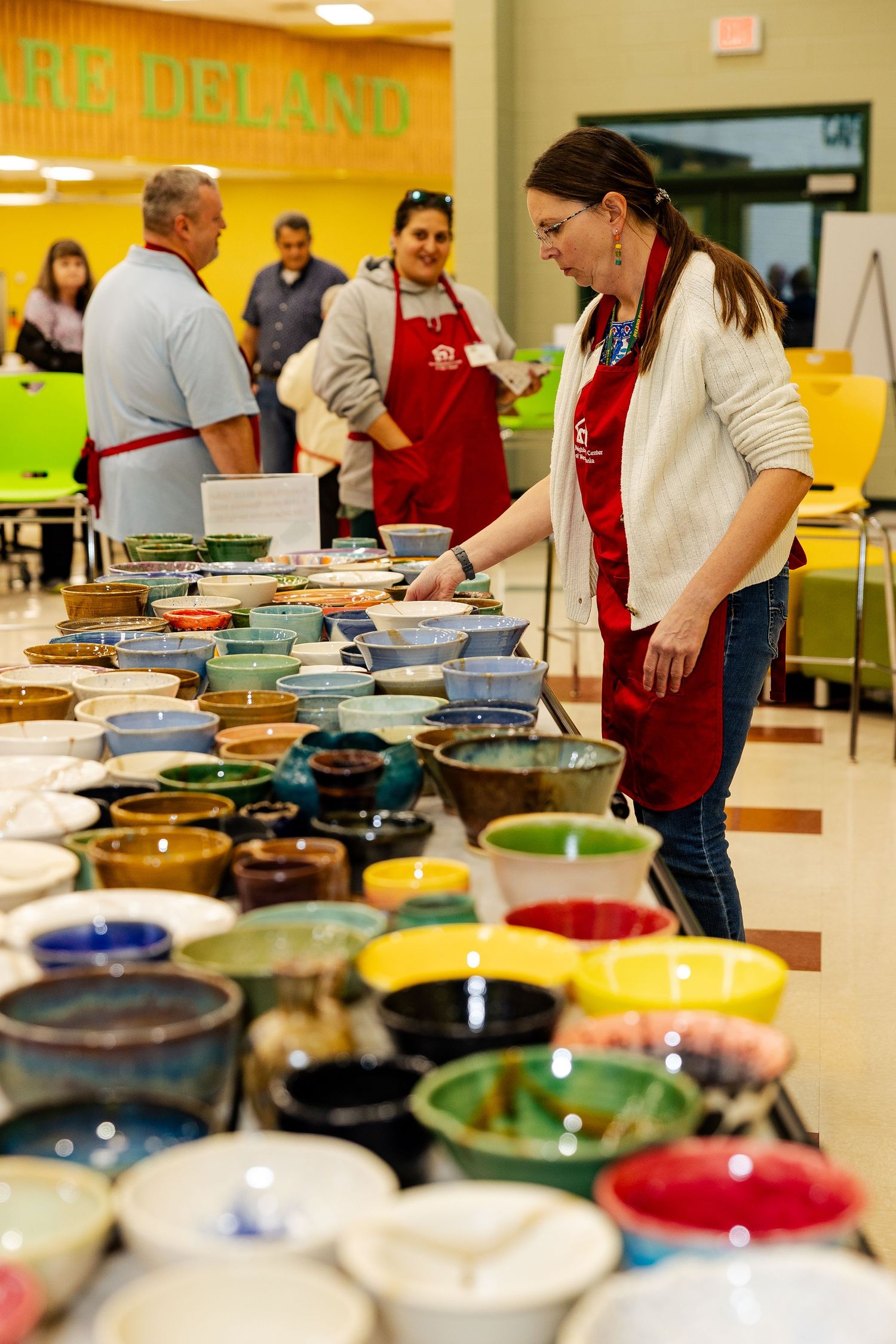 Volunteer in white sweater arranging colorful handmade ceramic bowls at a charity fundraiser.