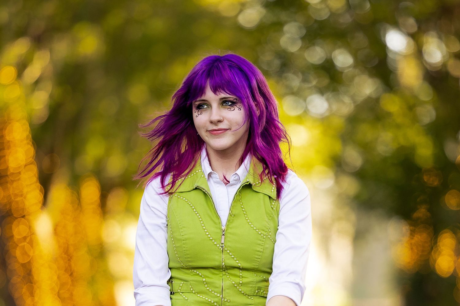 Young woman with vibrant purple hair wearing a green vest smiles outdoors with bokeh trees in background.