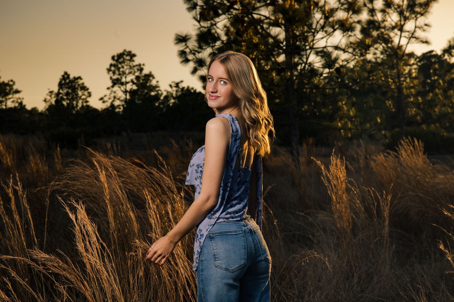 Young woman in jeans smiling over shoulder in golden field at sunset.
