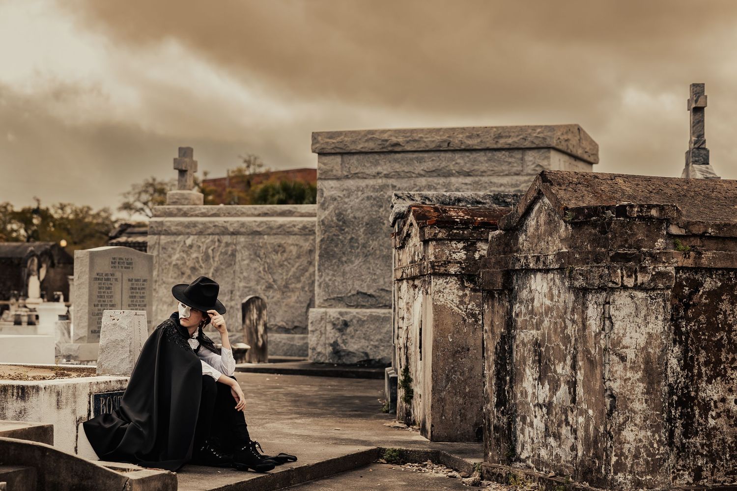 Person in black cape and wide-brim hat sitting contemplatively on tomb in historic cemetery.