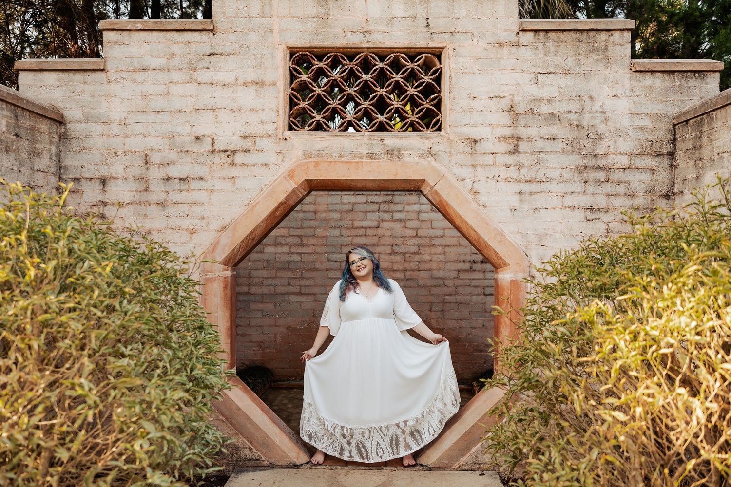 Woman in white dress posing in ornate octagonal garden archway surrounded by lush greenery and brick wall.
