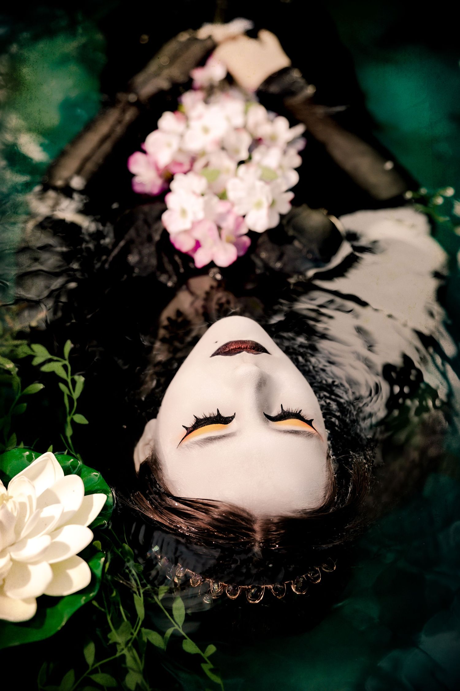 Woman in black floating in dark water surrounded by pink and white flowers, eyes closed.