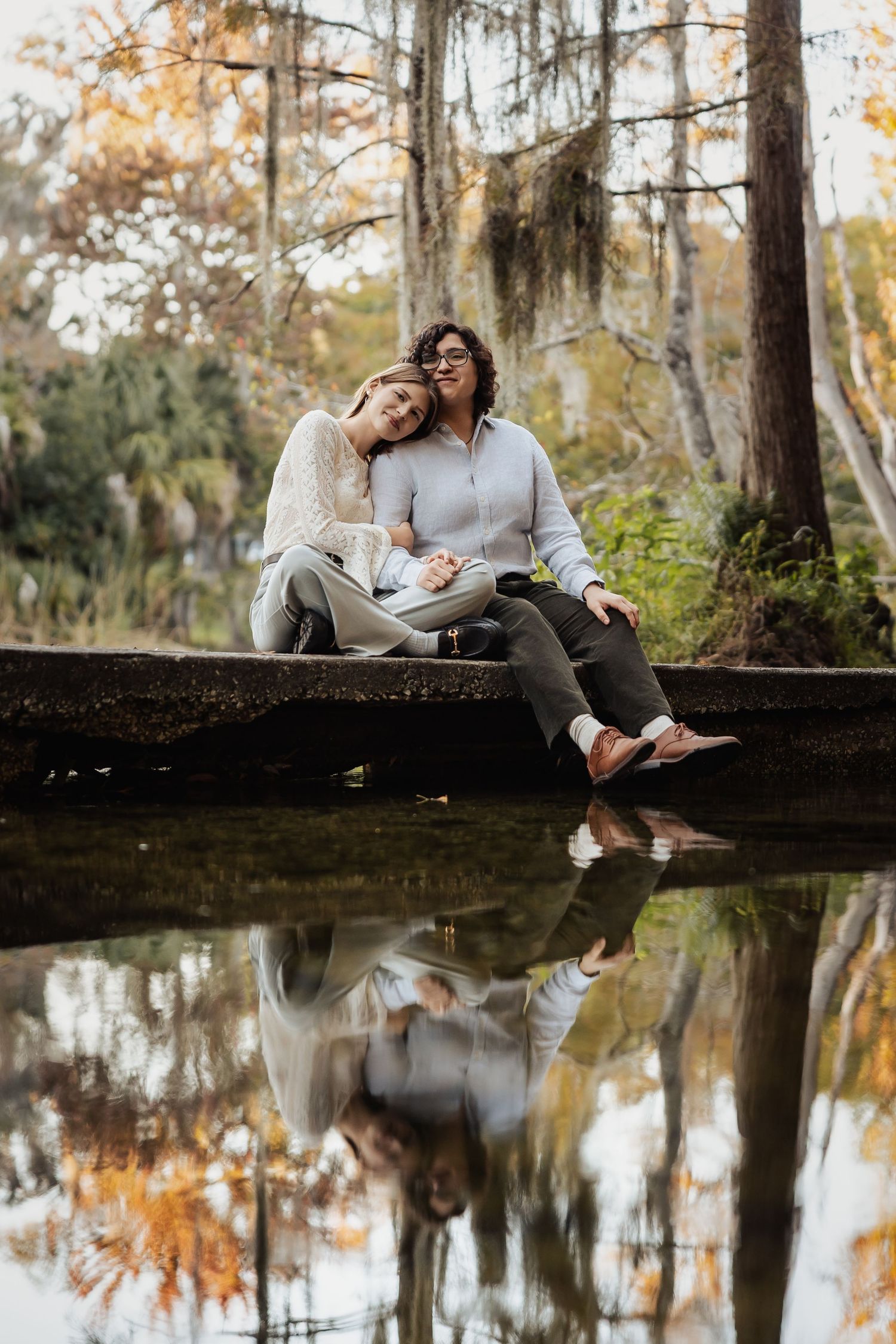 Two women sit closely together on a rock by a still pond, their reflections mirrored in the water.