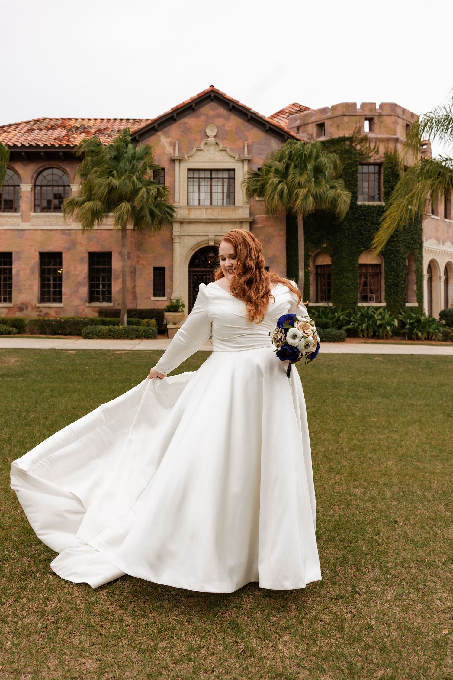 Redhead bride twirls white long-sleeve wedding gown on lawn before Spanish-style mansion.