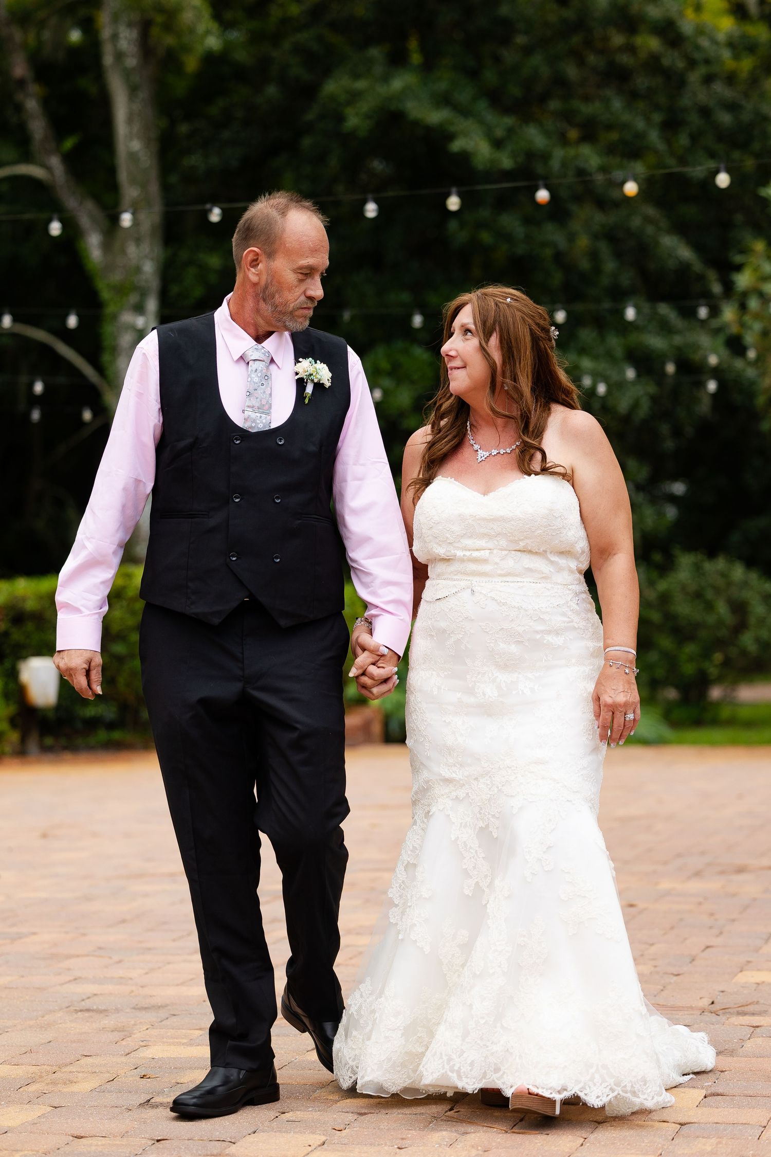 Newlyweds walk hand in hand on brick patio with string lights and lush greenery in background.