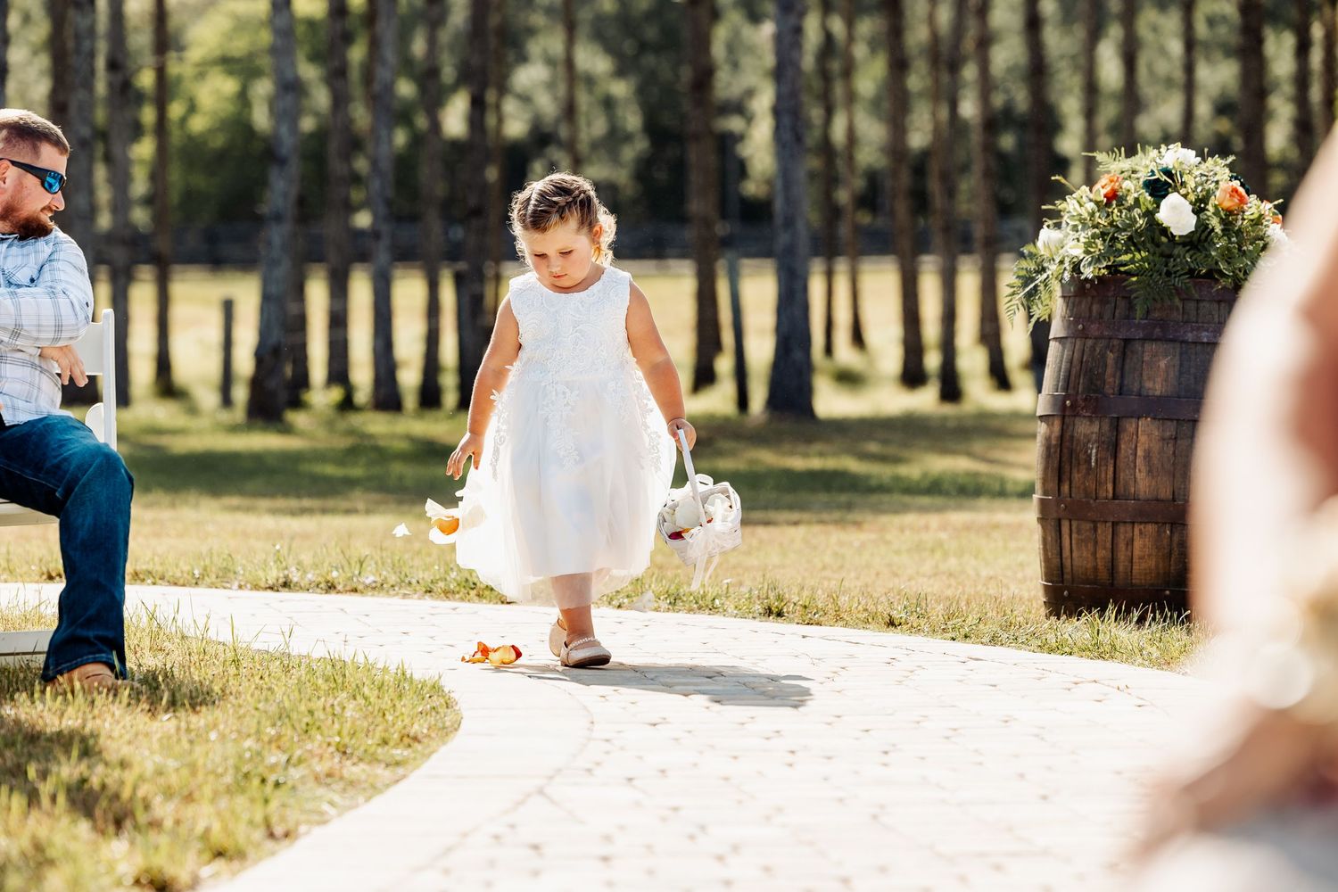Young flower girl walking down outdoor ceremony aisle near rustic barrel decor.