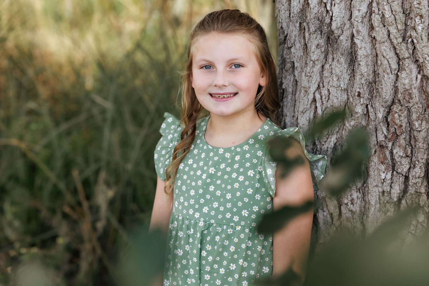 Child in polka dot green dress poses beside tree trunk.
