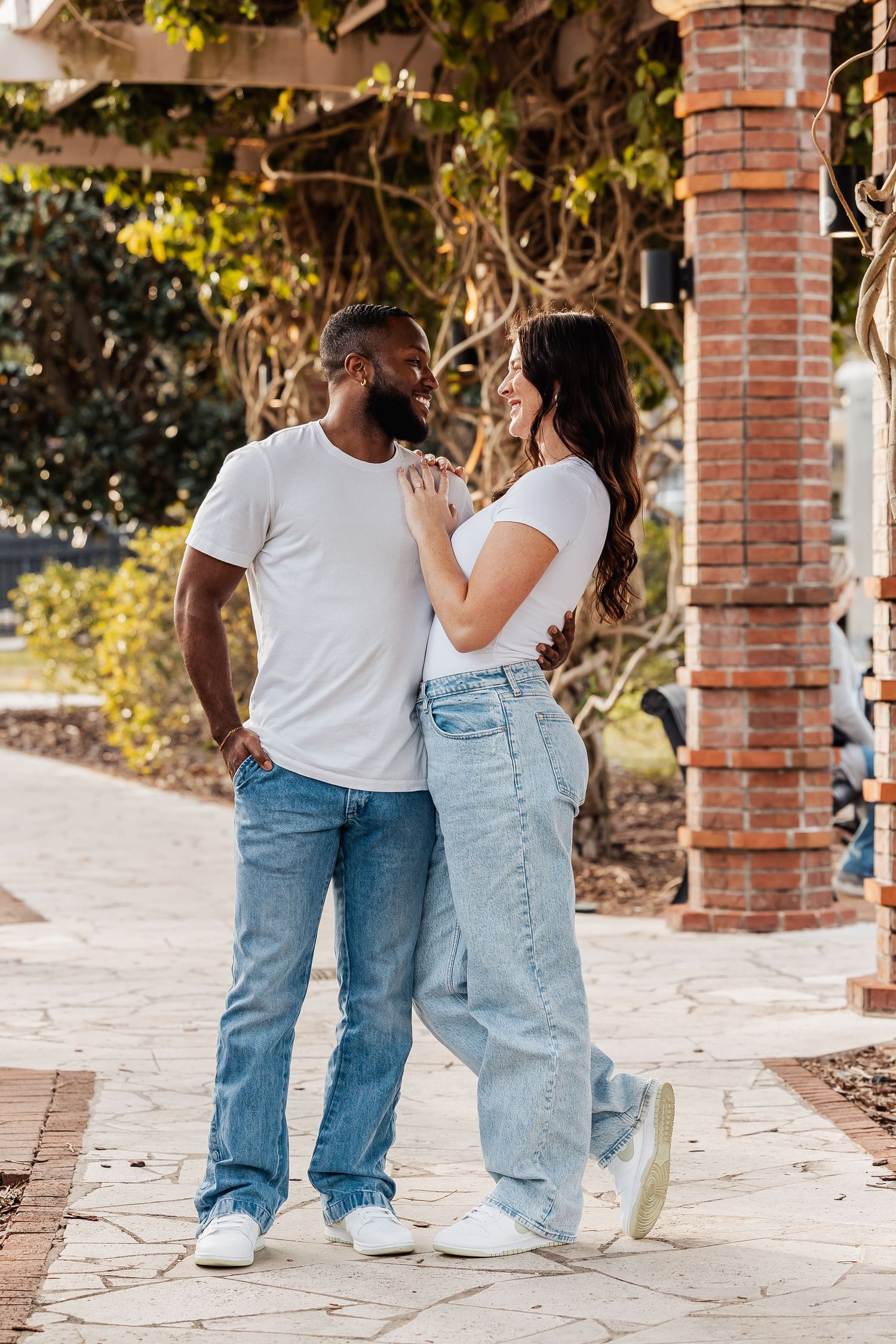A couple shares an intimate moment while wearing matching white t-shirts and jeans beside a brick column.