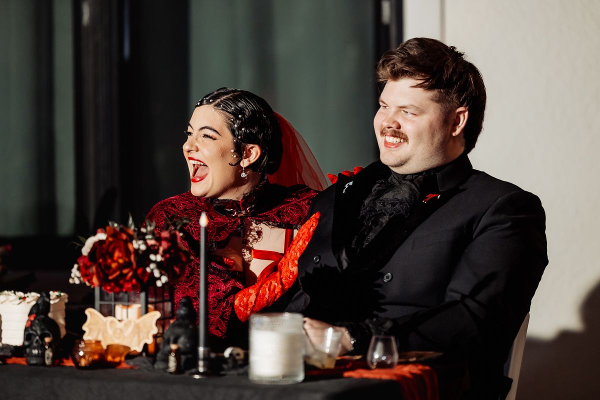 Two people laugh together at a formal event, one wearing red and one in black, with red floral decorations on the table.