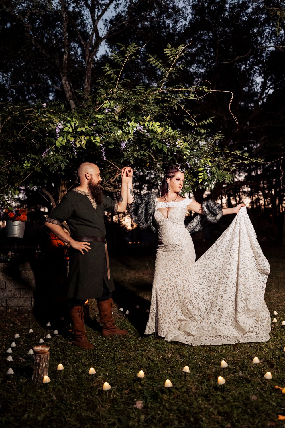 A couple poses dramatically in a garden setting with string lights, featuring a flowing lace dress and medieval attire.