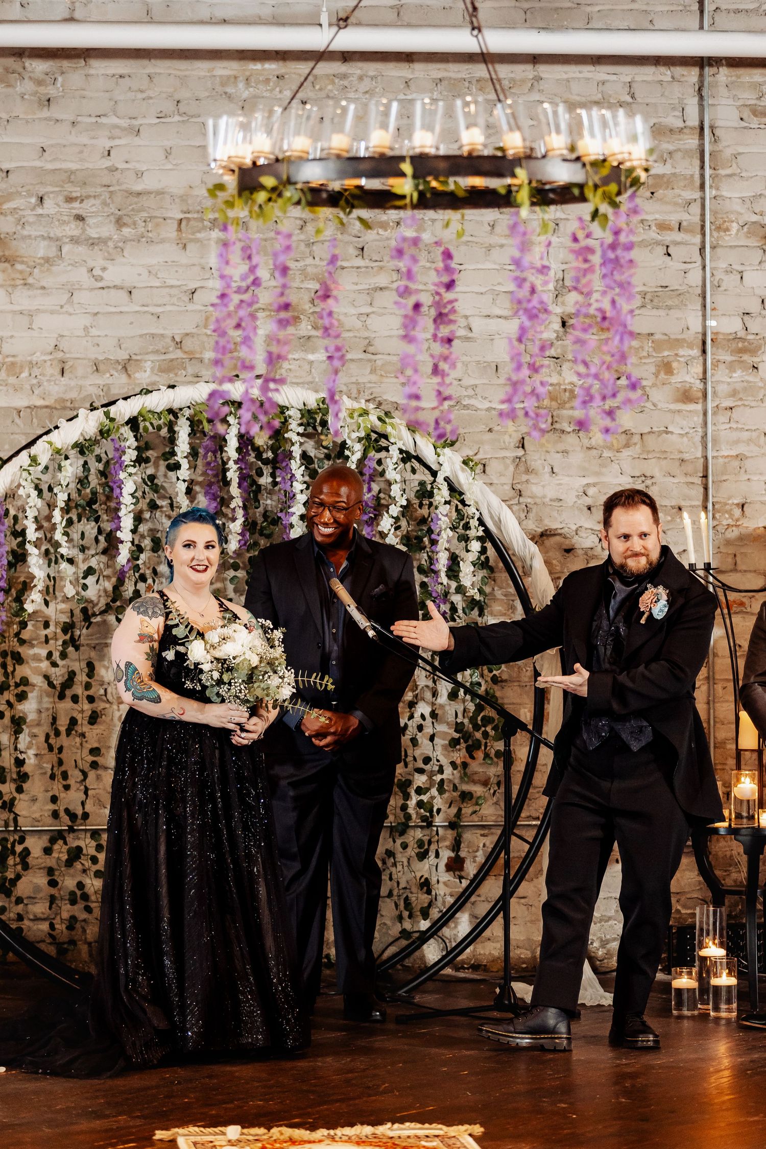Bride and groom in matching black attire stand with officiant at a circular floral wedding arch.