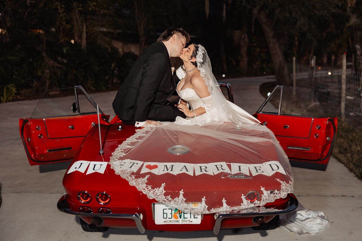 Newlyweds share a kiss on a classic red Corvette convertible with Just Married sign at night.