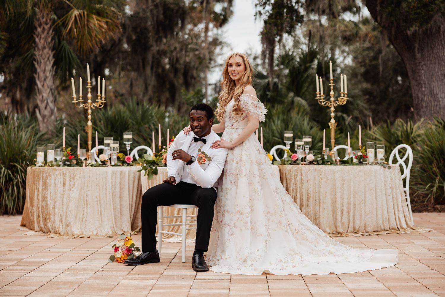Interracial couple posing elegantly at outdoor wedding reception with gold candelabras and floral arrangements.