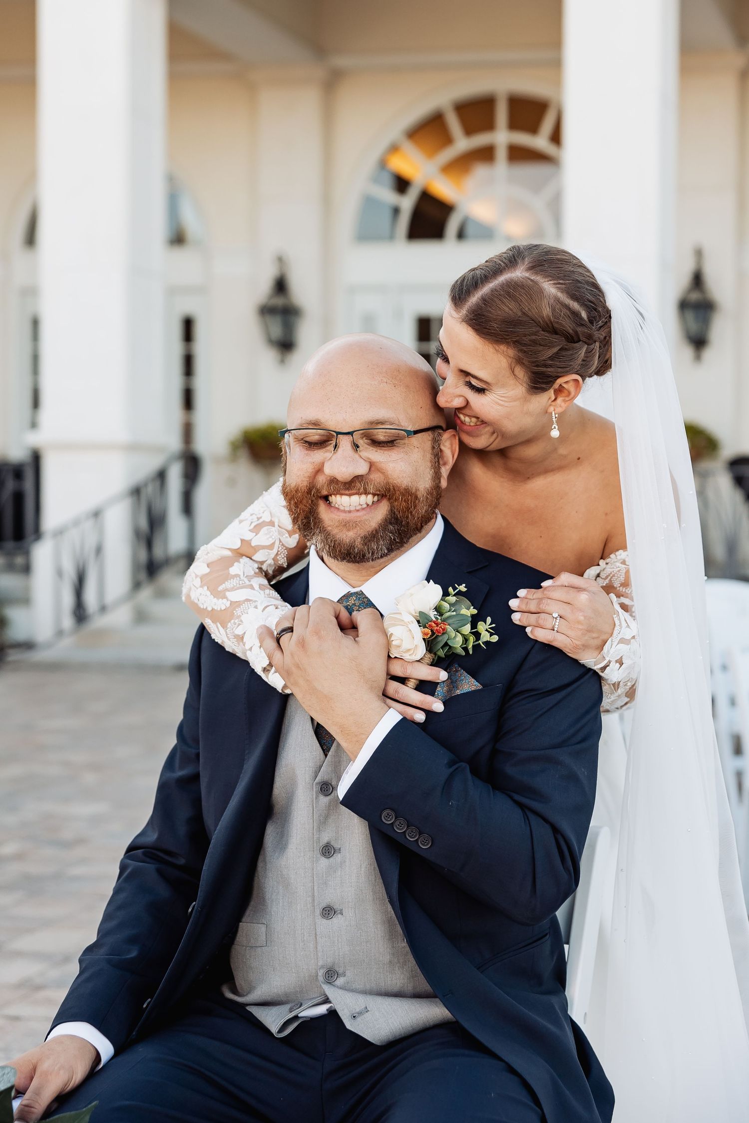 Smiling groom in navy suit embraced by bride in lace wedding dress on venue steps.