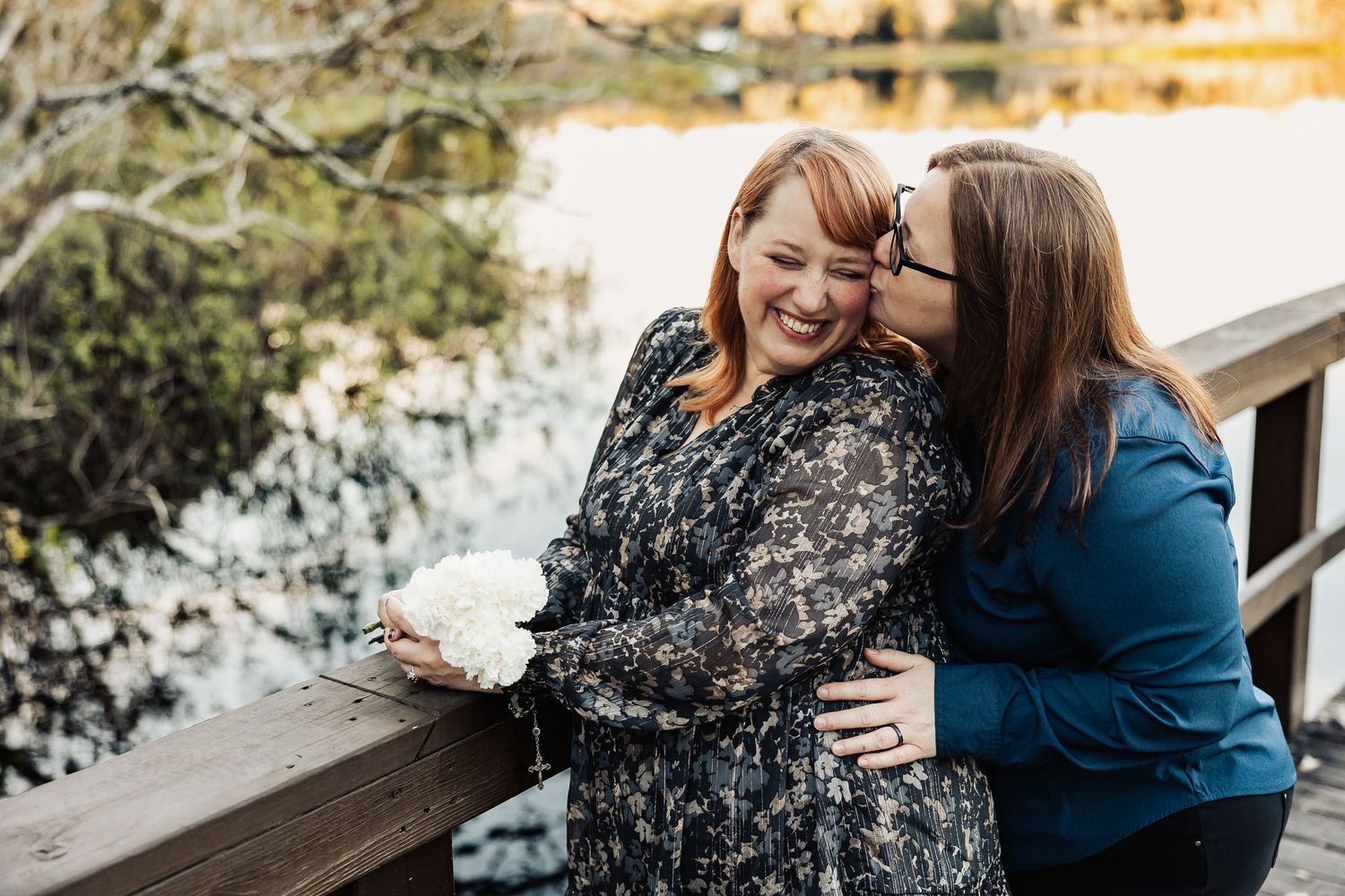 A woman kisses her smiling partner on the cheek on a wooden bridge, holding white flowers near a serene lake.