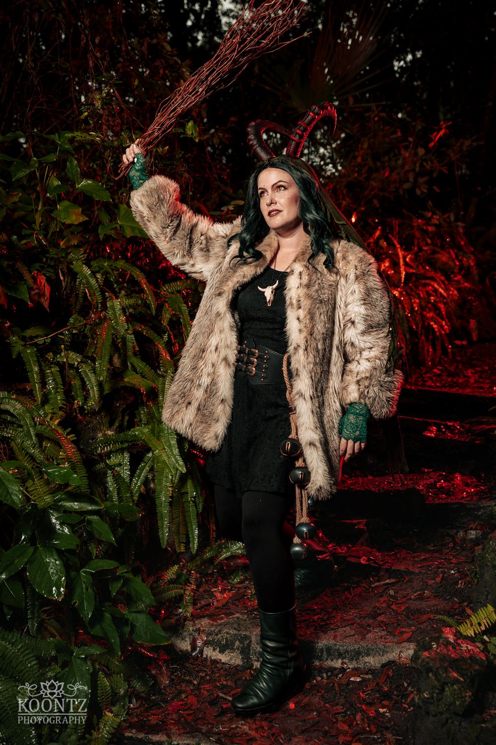Woman in fur coat with horns and broom poses dramatically in dark forest setting for Halloween cosplay photography.