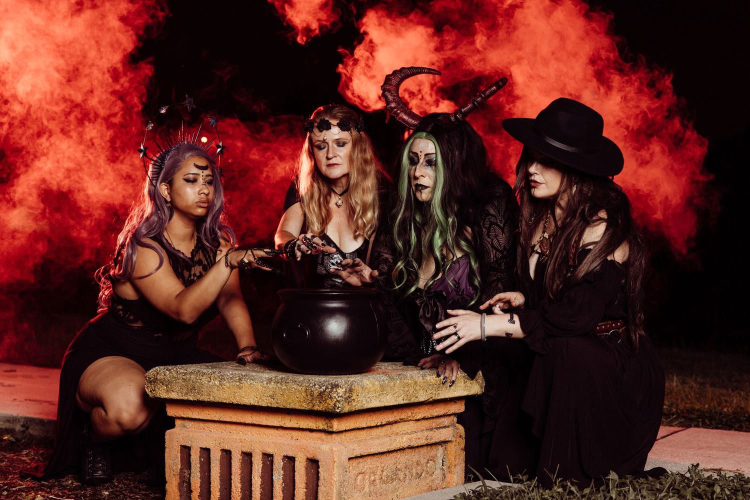 Four women dressed as witches pose around a stone cauldron with eerie red smoke backdrop at night.