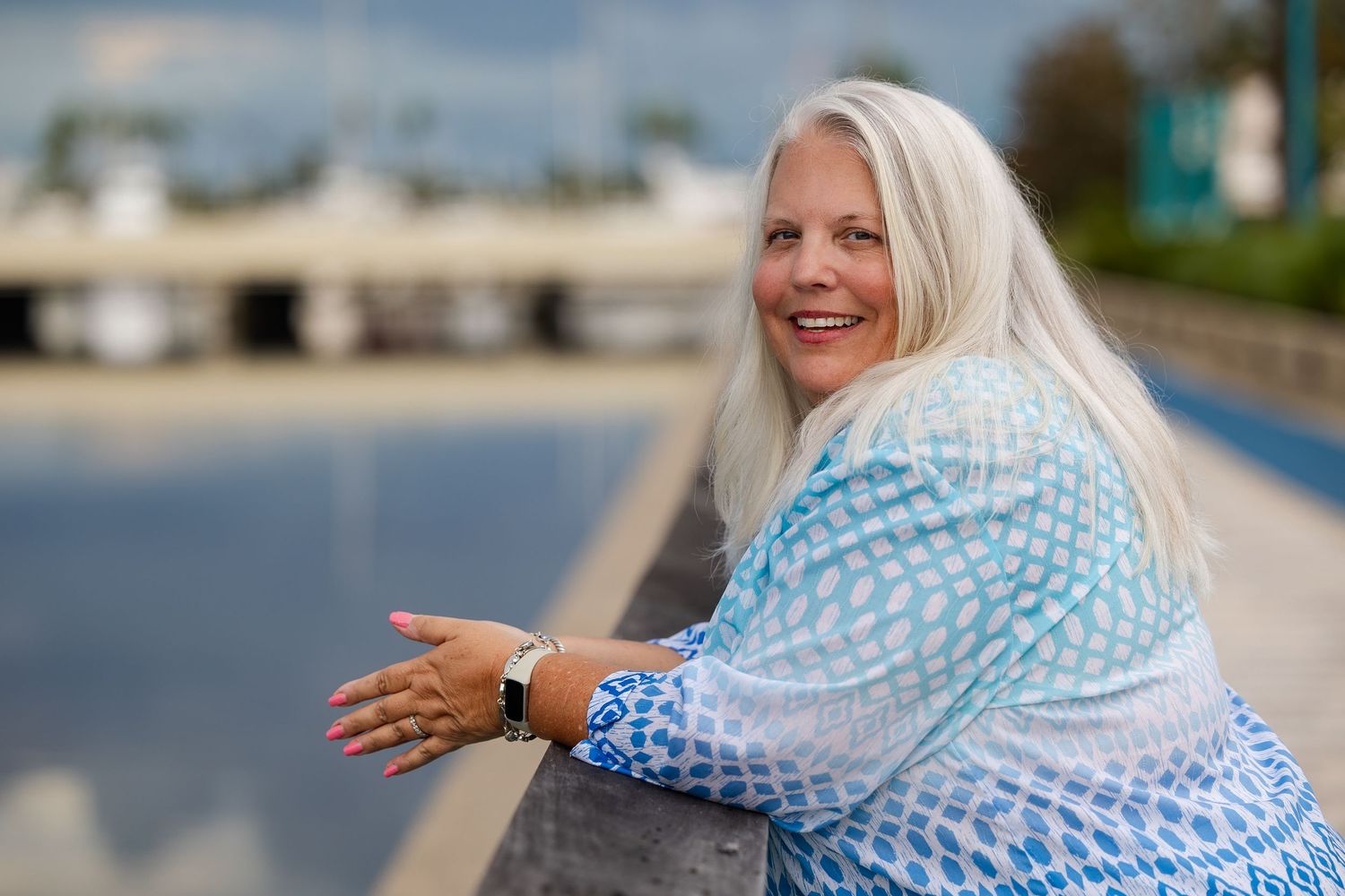 Smiling woman with long gray hair leaning on a railing near a waterfront on a sunny day.