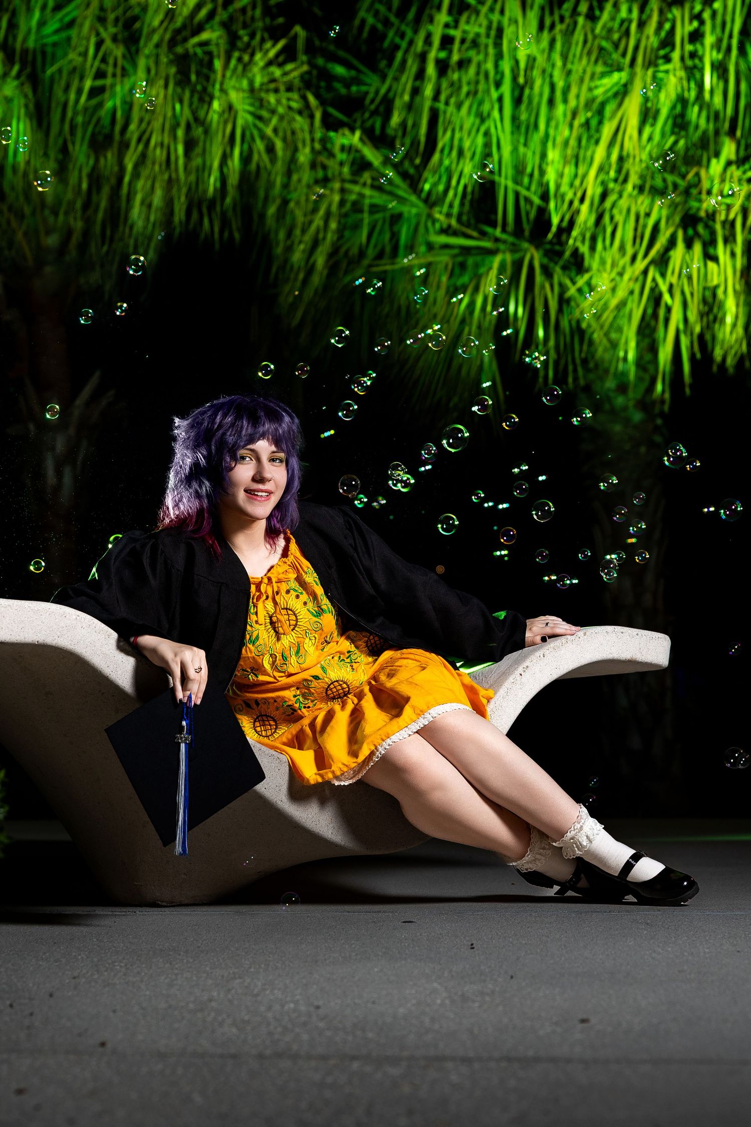 A young woman in an orange dress and graduation gown sits on a white bench surrounded by bubbles and green-lit palms.