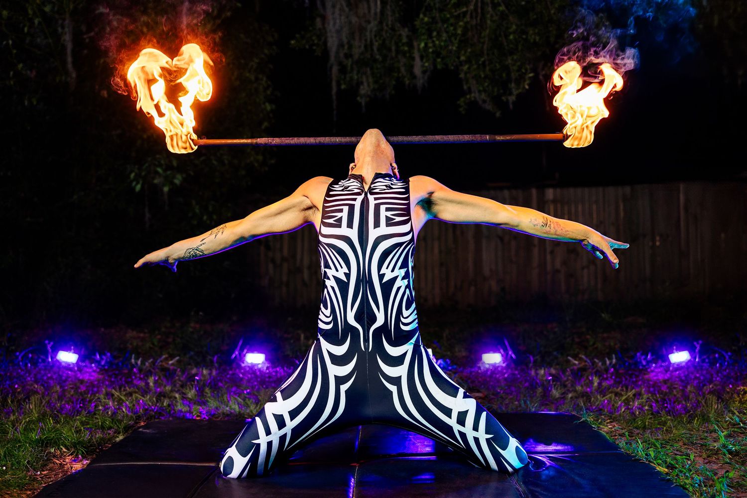Aerial fire performer in tribal bodysuit poses with two flaming torches at night.