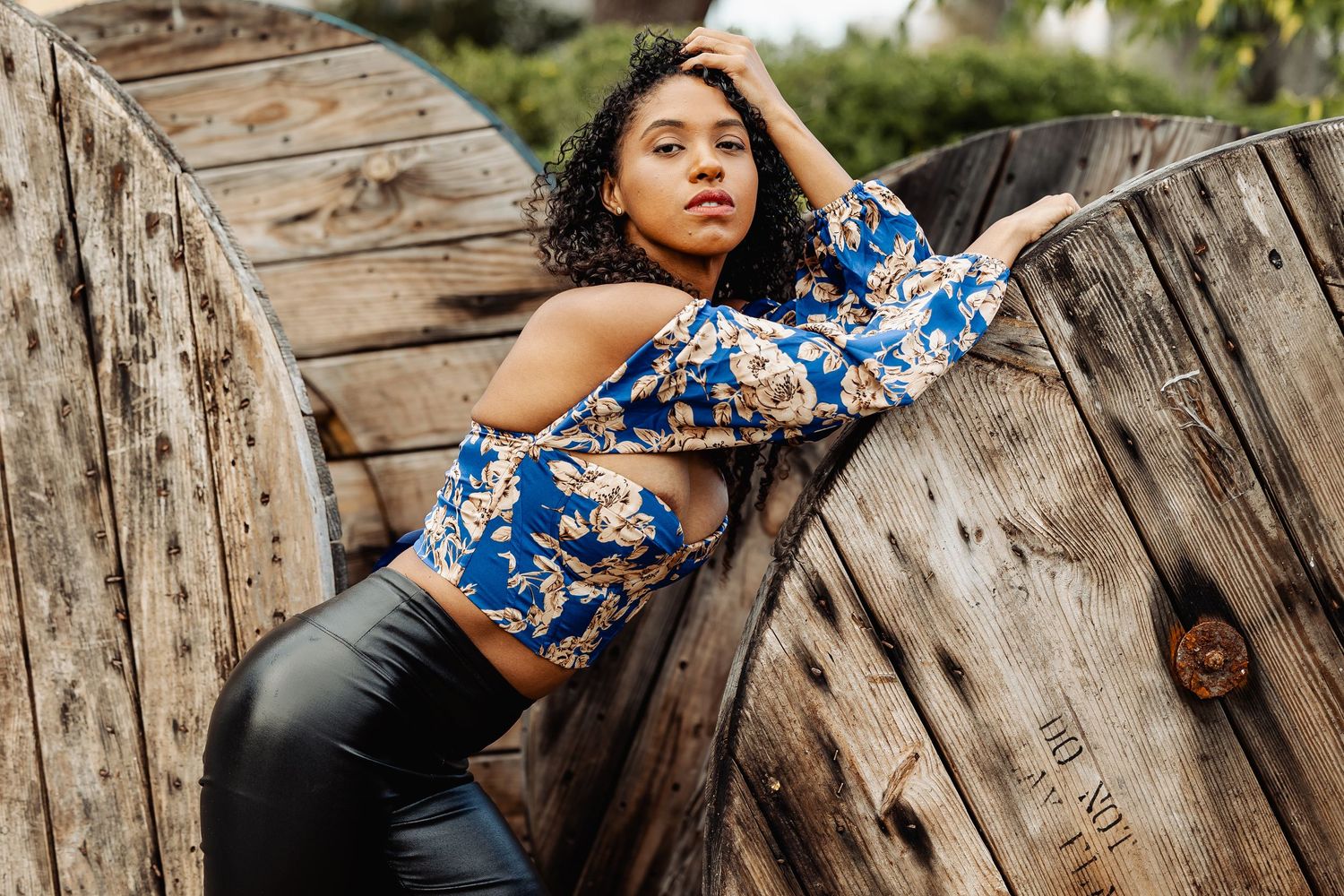 Woman in blue floral crop top and black leather pants leans against a large wooden spool.