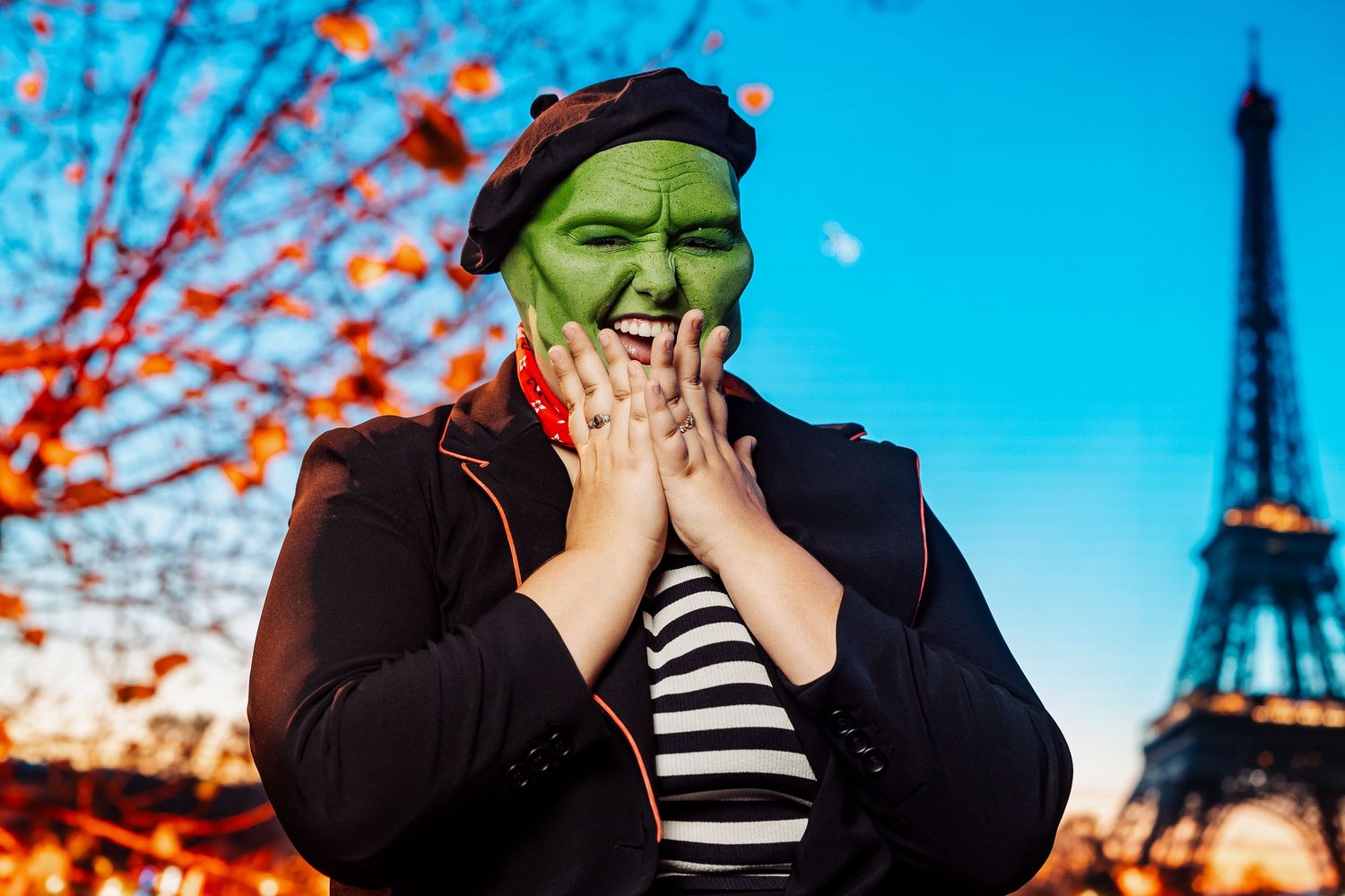 Person wearing green face paint Mask costume with beret and striped shirt posing in front of the Eiffel Tower in Paris.