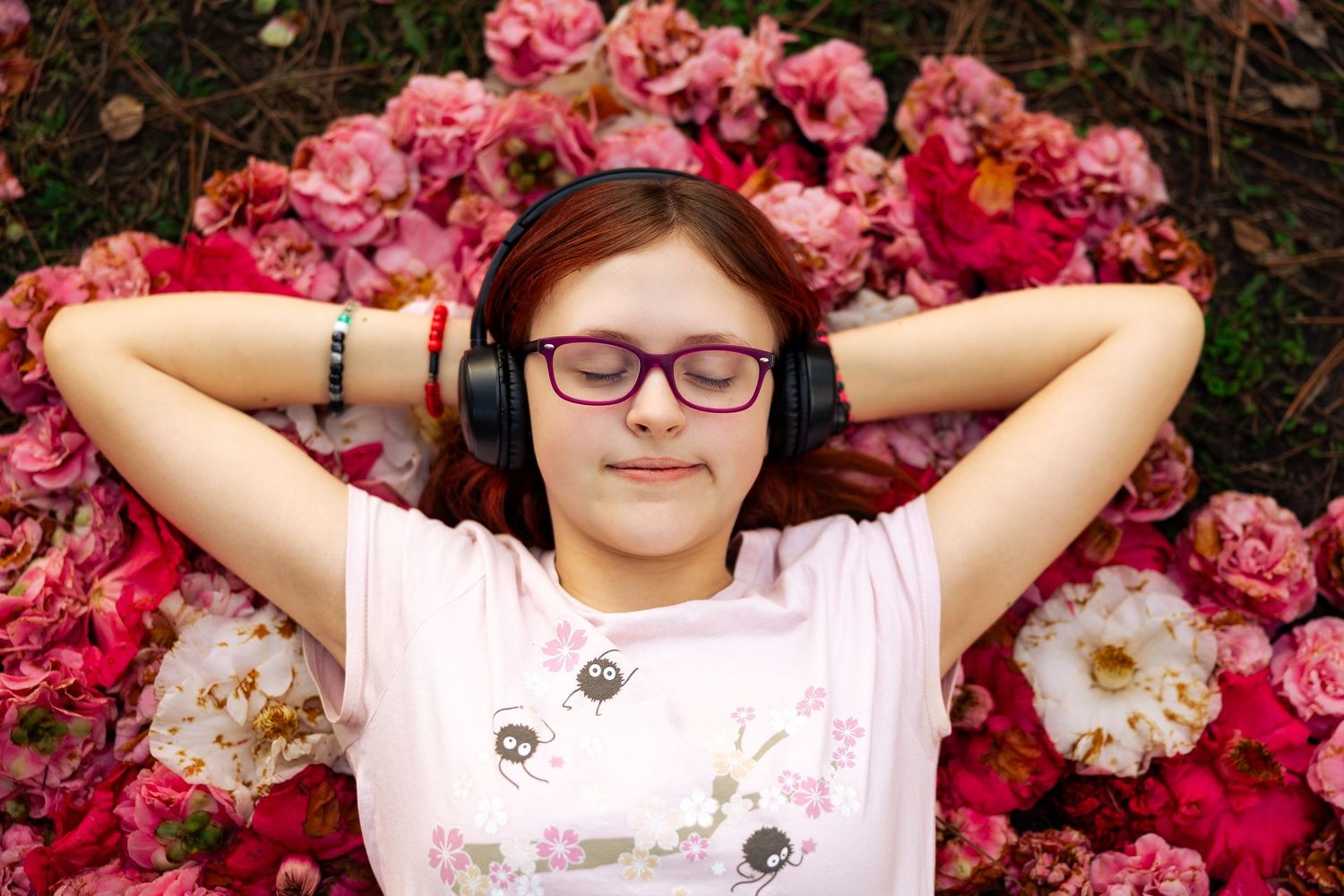 Young girl with glasses and headphones relaxing on a bed of pink flowers with eyes closed.