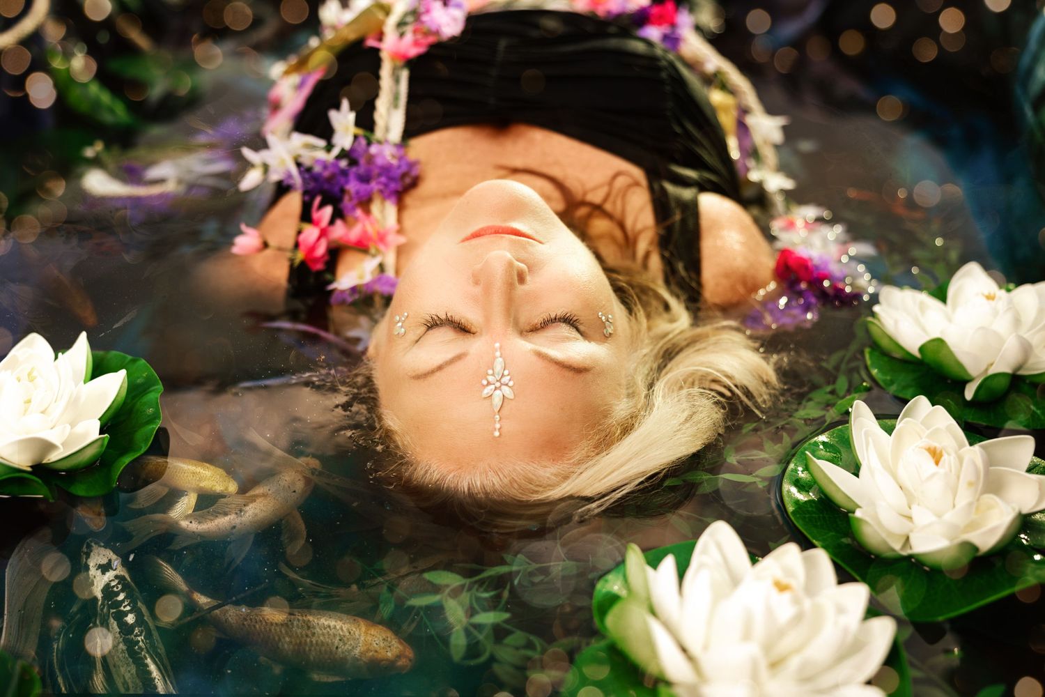 A woman floats serenely in a koi pond surrounded by white lotus flowers with a crystal gem on her forehead.