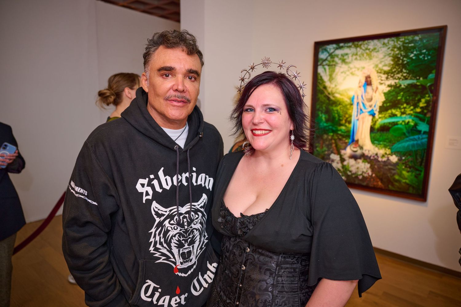 People gather at an art gallery opening event with colorful paintings displayed on white walls in the background.